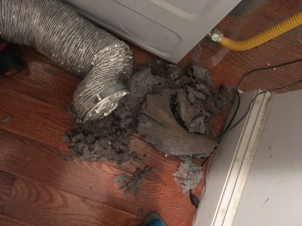 Dryer vent with large pile of lint on floor. Silver vent tubing, white appliance, and yellow gas line.