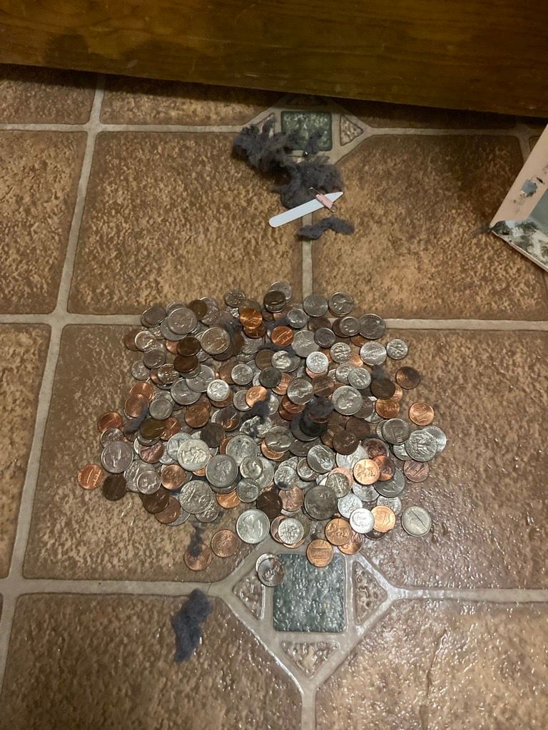 Pile of coins on a brown tiled floor, with a bit of crumpled paper and a small scrap of fabric.