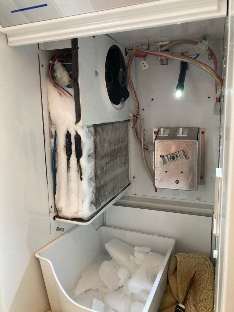 Interior of a refrigerator showing a frozen evaporator coil, fan, ice bucket, and wiring.