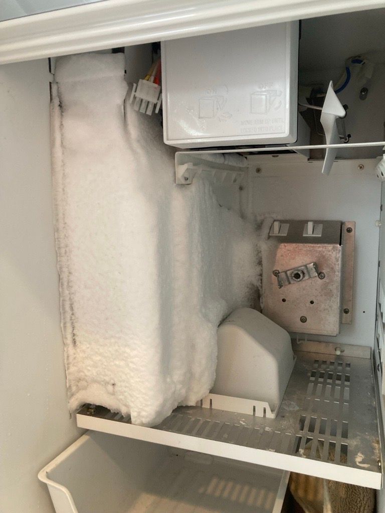 Interior of a freezer with heavy frost buildup on the back wall and components; an ice maker and shelf are visible.
