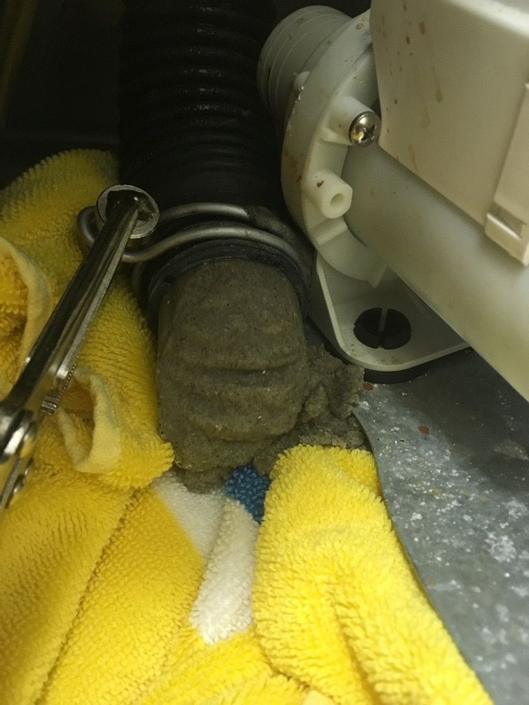 Wrench removing a hose from a washing machine pump; gray debris is visible around the connection. Yellow towels are beneath.