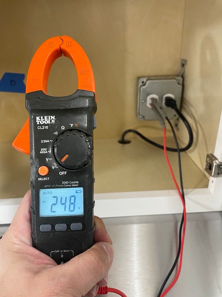 A person holds a clamp meter, reading 24.8, near an electrical outlet. Wires are plugged in.