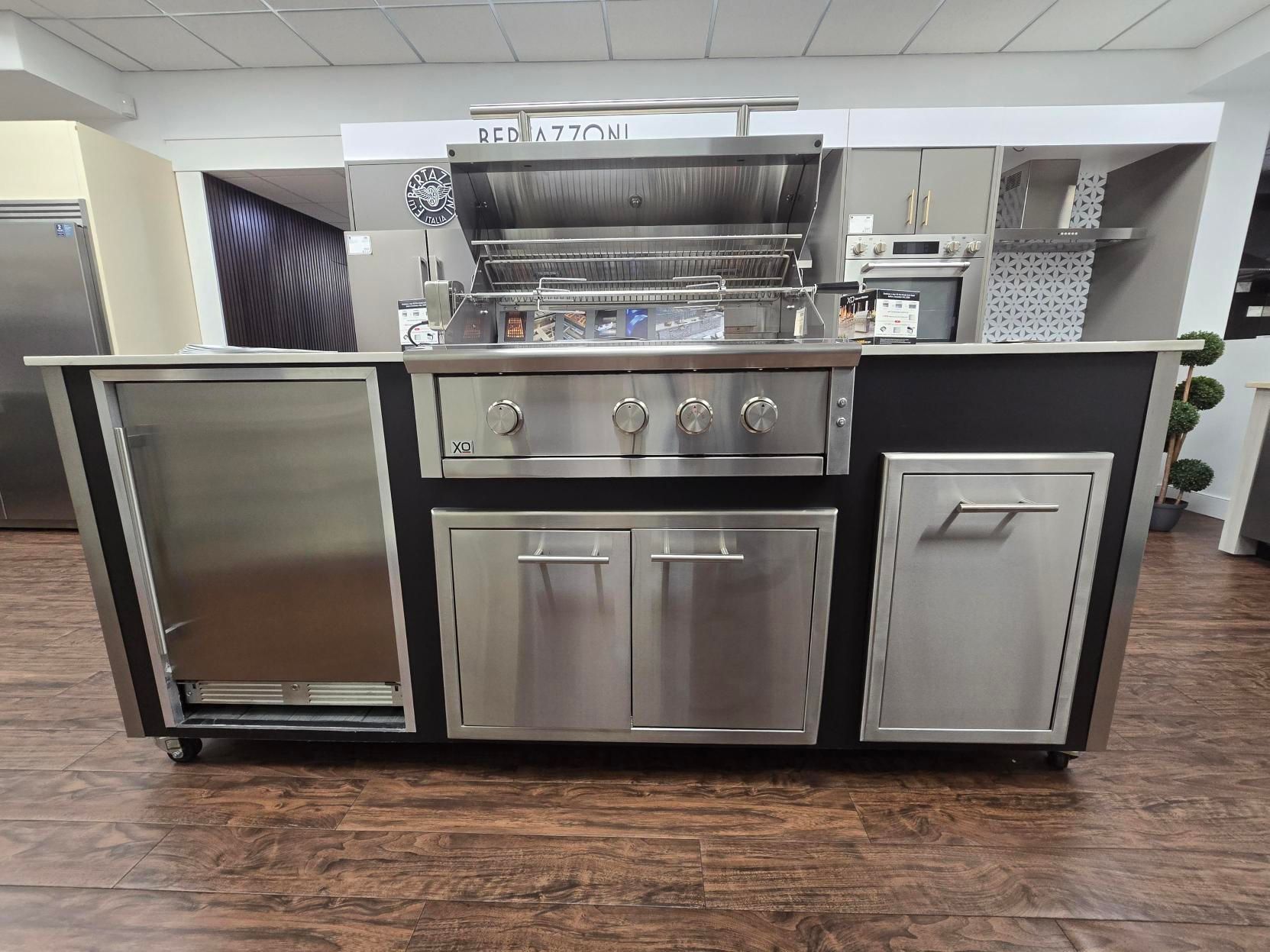 Stainless steel outdoor kitchen with grill, fridge, and storage cabinets, in a showroom.