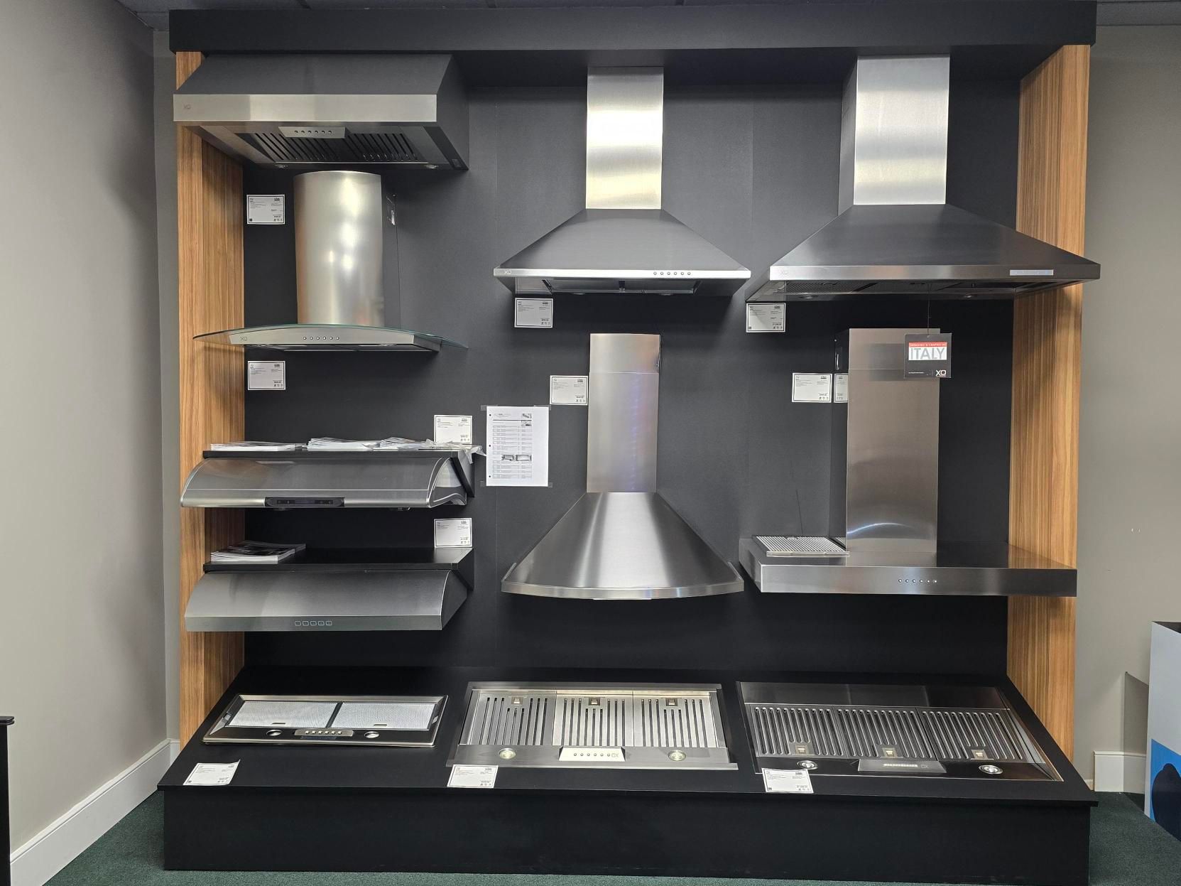 Display of stainless steel range hoods in a showroom against a black backdrop, with wood accents.