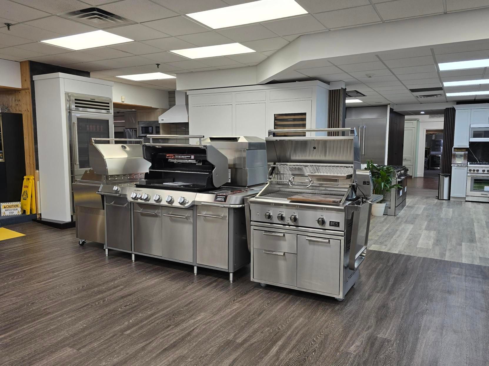 Indoor showroom displays stainless steel grills and appliances on wooden floor.