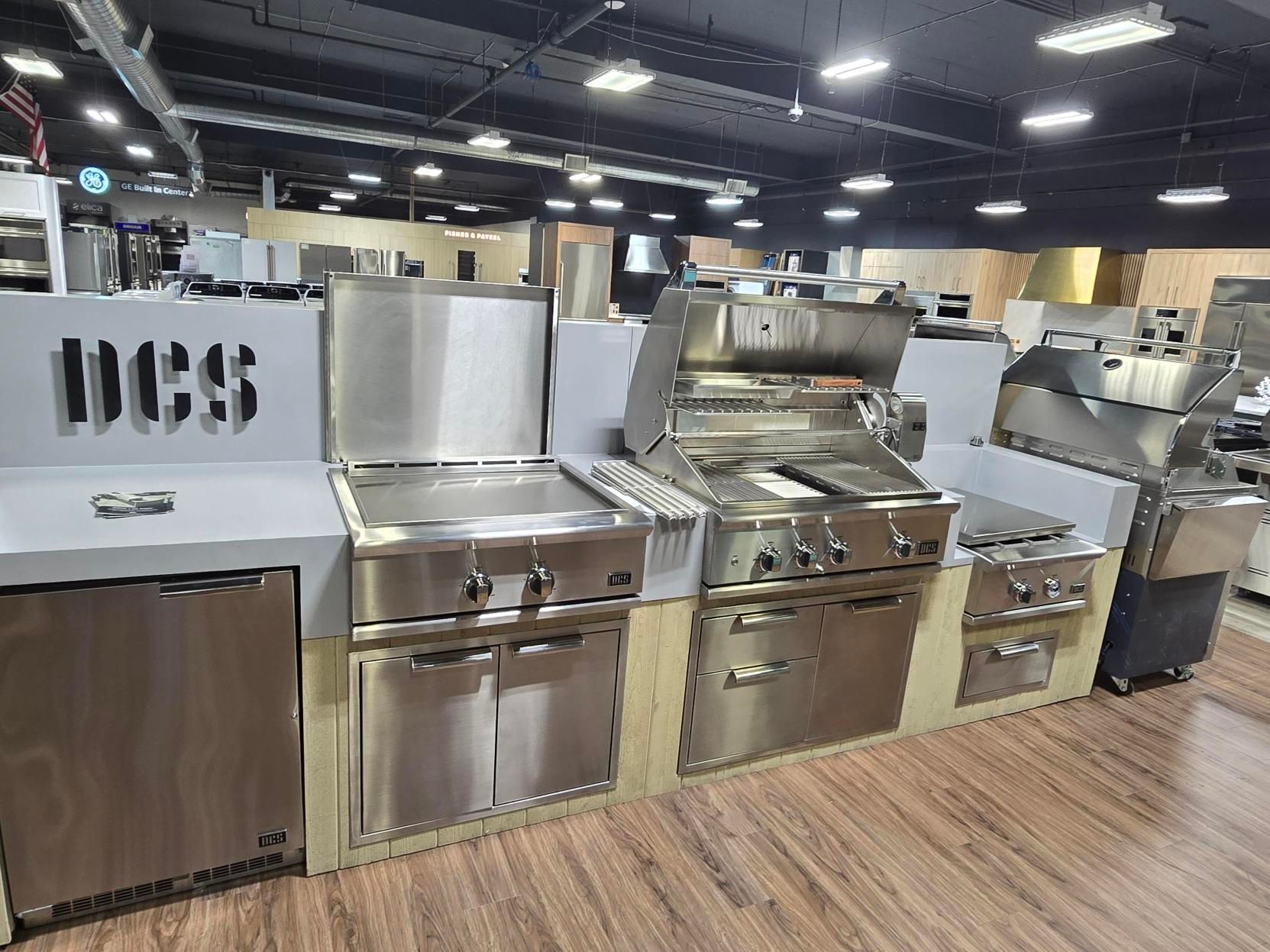 A showroom display of stainless steel DCS outdoor kitchen appliances, including grills and refrigeration, on wooden platforms.