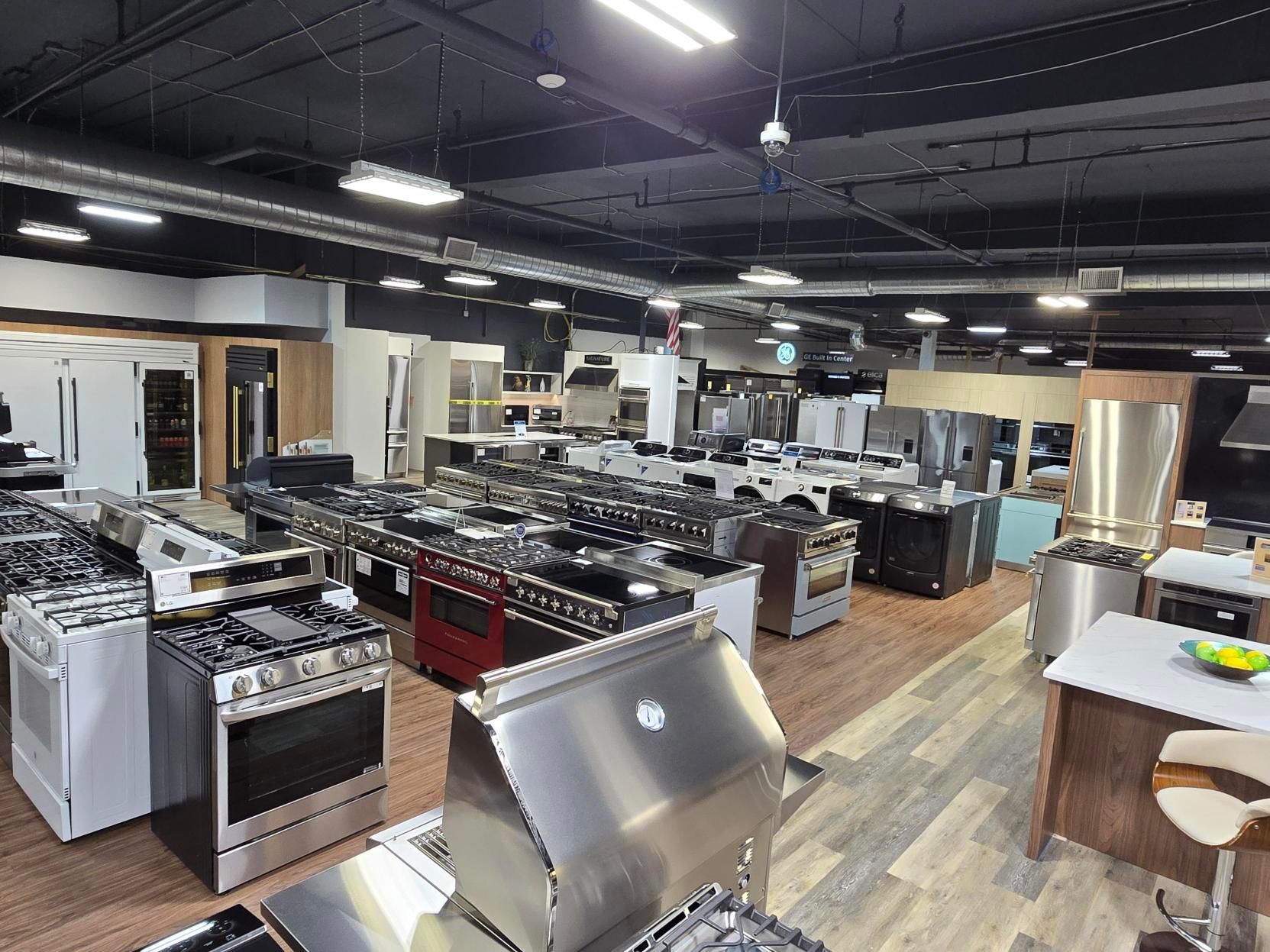 Appliances showroom with stoves, refrigerators, and other kitchen equipment displayed under bright lighting.