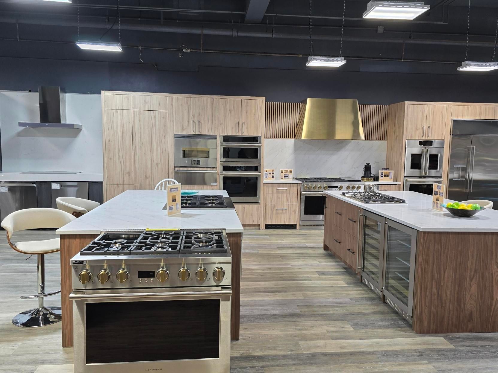 A showroom kitchen displays various cabinets and appliances with two island cooktops and wood accents.