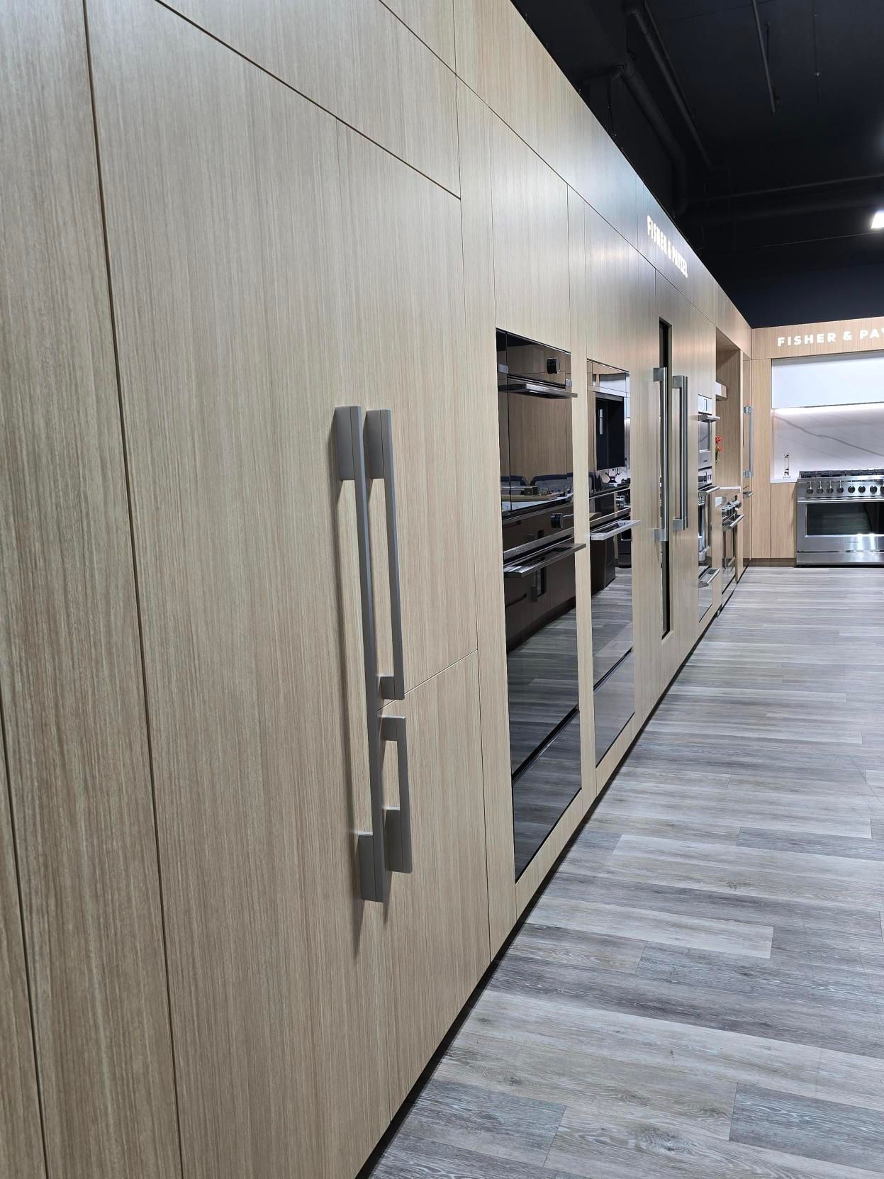 Wooden paneling with recessed appliances on display in a showroom, silver handles, and gray flooring.