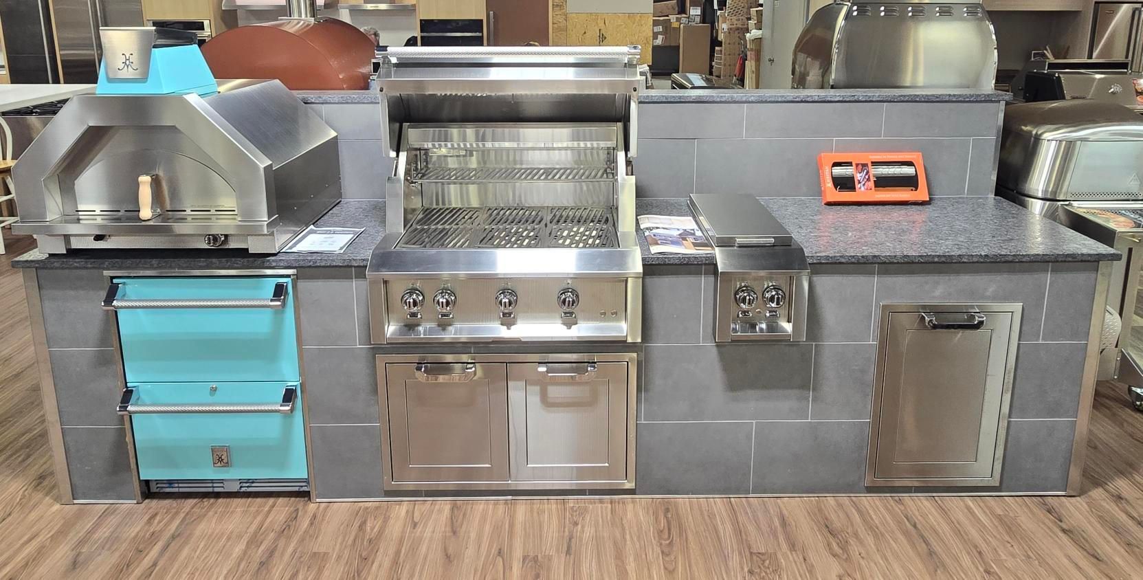 Outdoor kitchen island with pizza oven, grill, side burner, storage, and a teal accent.