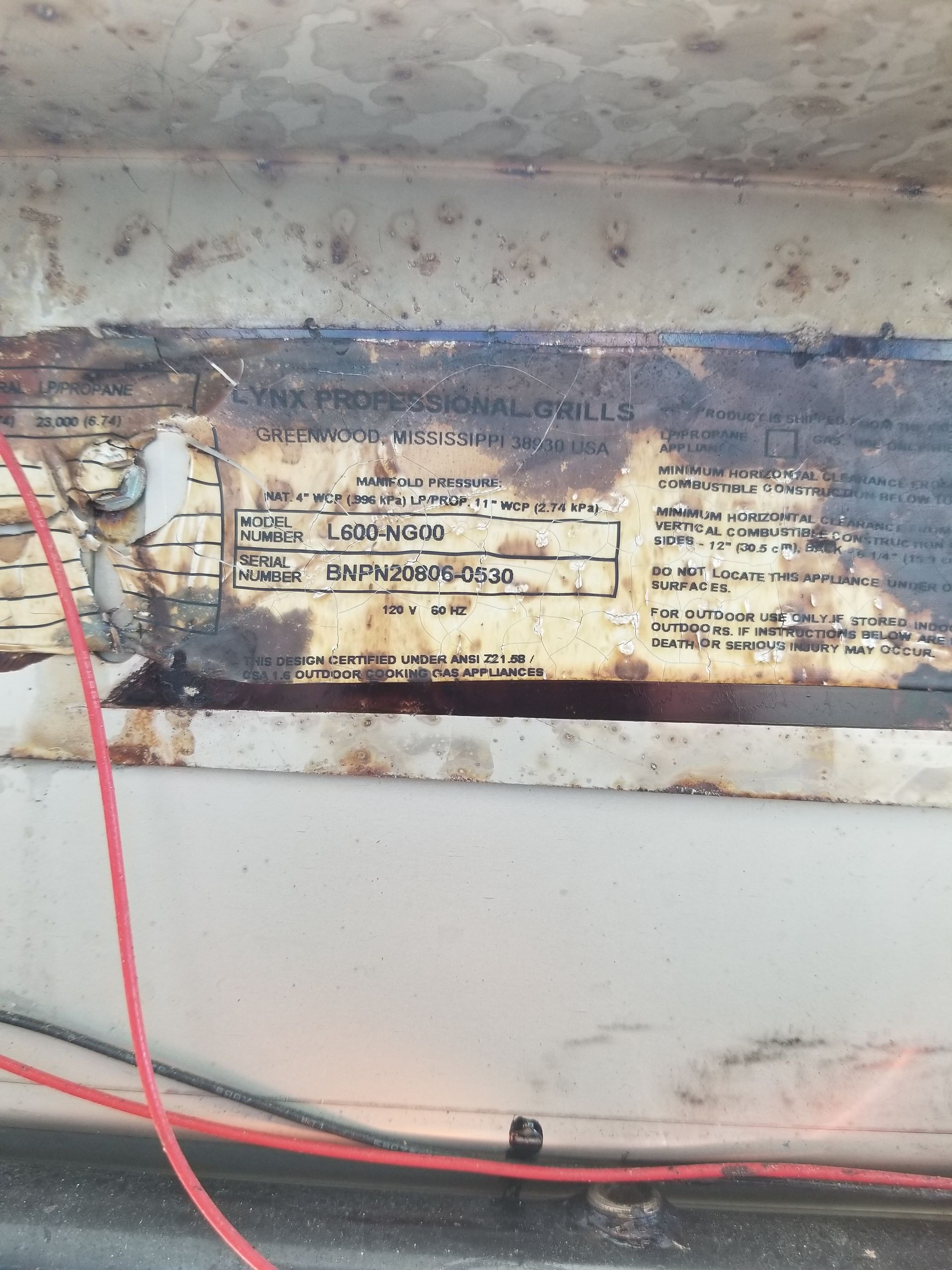 Rust-covered equipment information plate with faded text, mounted on a white surface. Red and black wires cross the bottom.