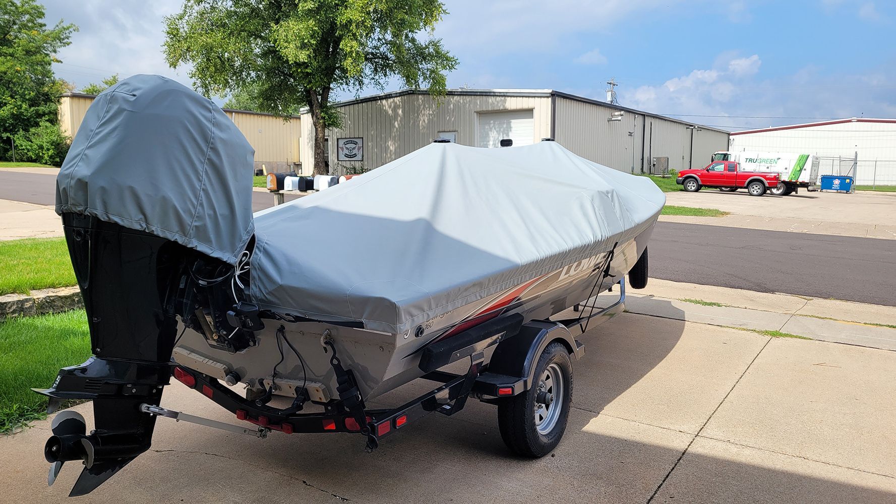 A boat covered with a gray tarp sits on a trailer in front of a building on a sunny day.
