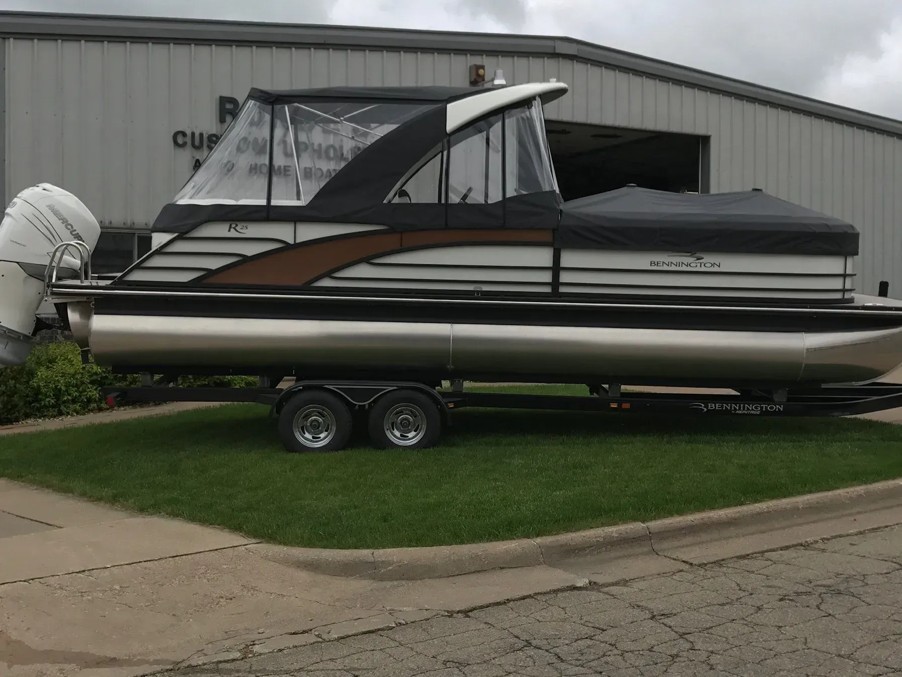 Pontoon boat with black canopy and silver pontoons, parked on grass in front of a building.
