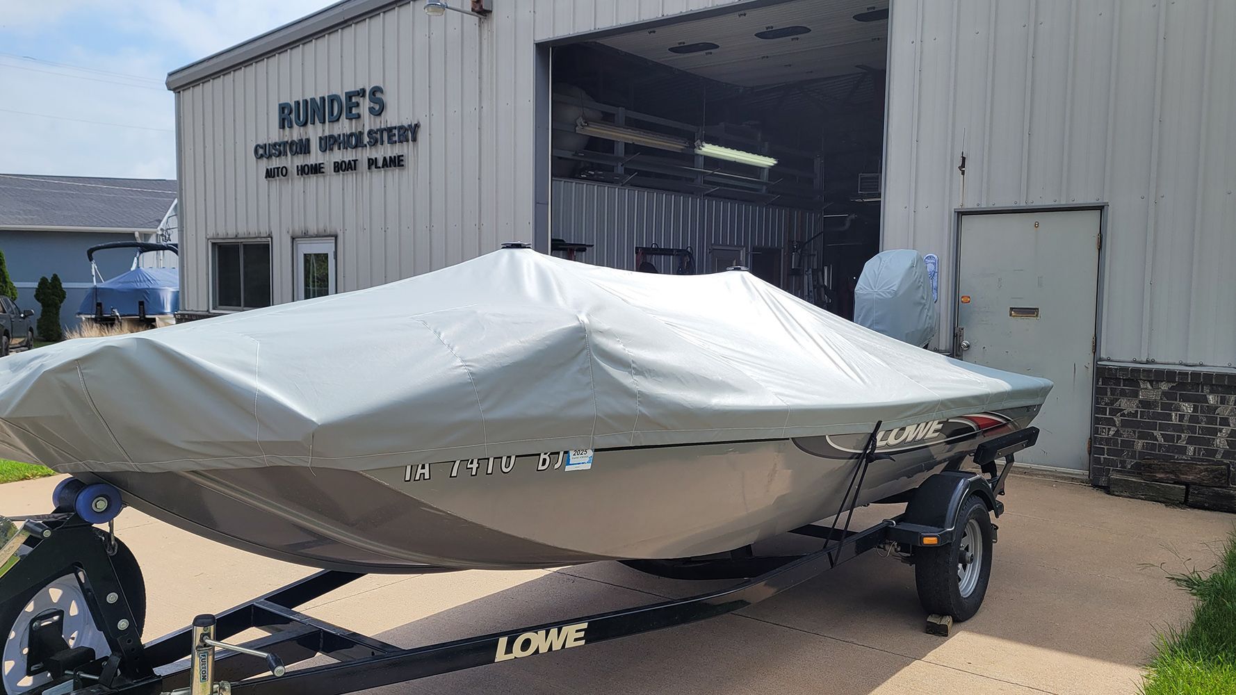 A covered Lowe boat on a trailer parked outside a building labeled "Burde's Custom Upholstery."