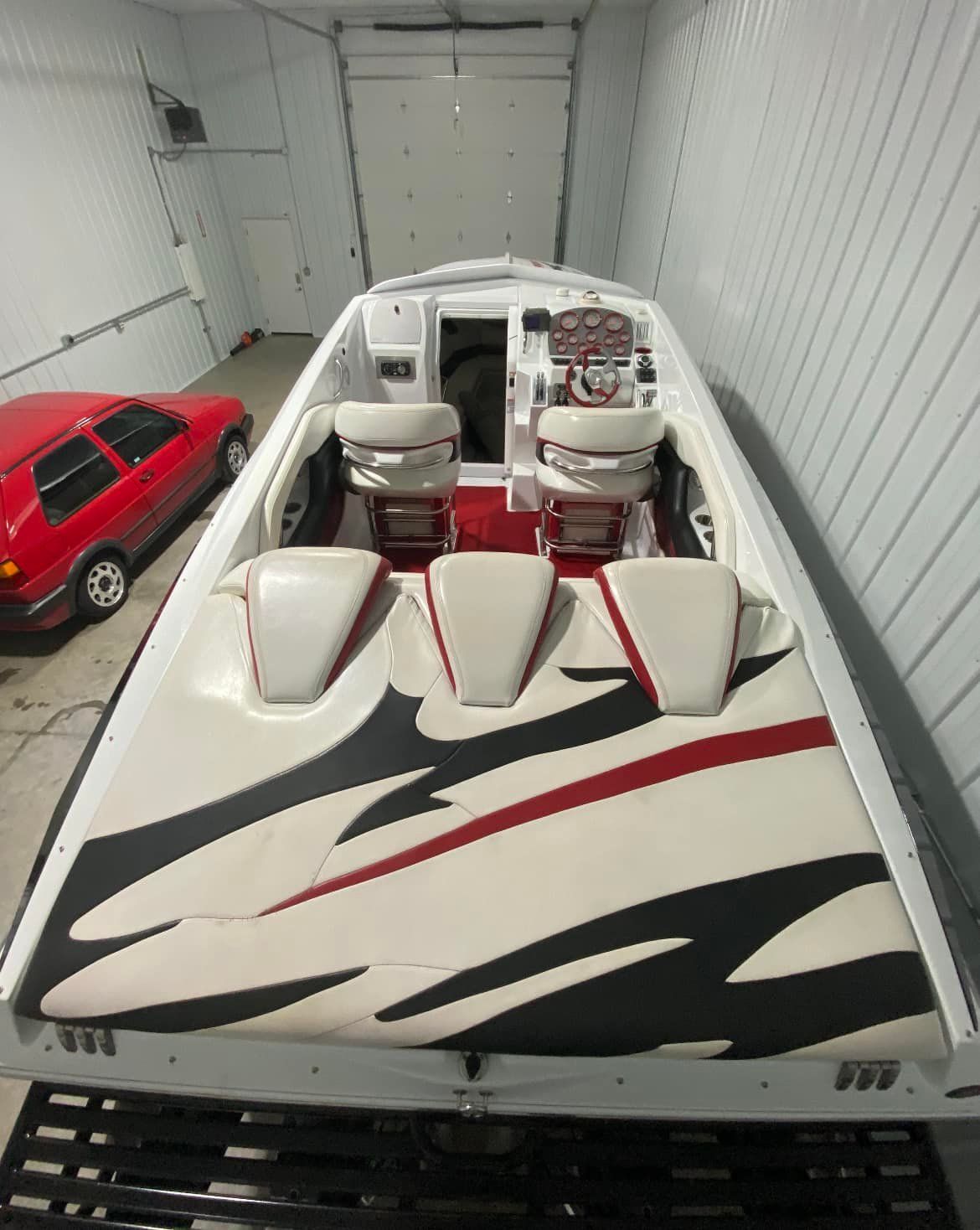 A white speedboat with red and black accents, red seats, and a red car parked nearby inside a building.