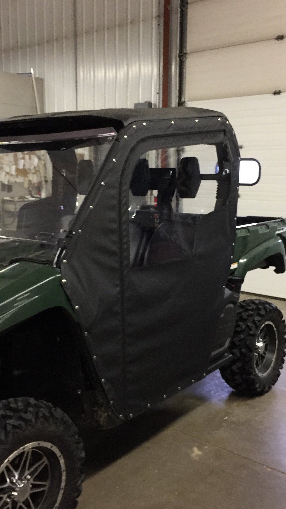 Dark green side-by-side with black fabric doors and roof, black side mirrors, and large off-road tires.