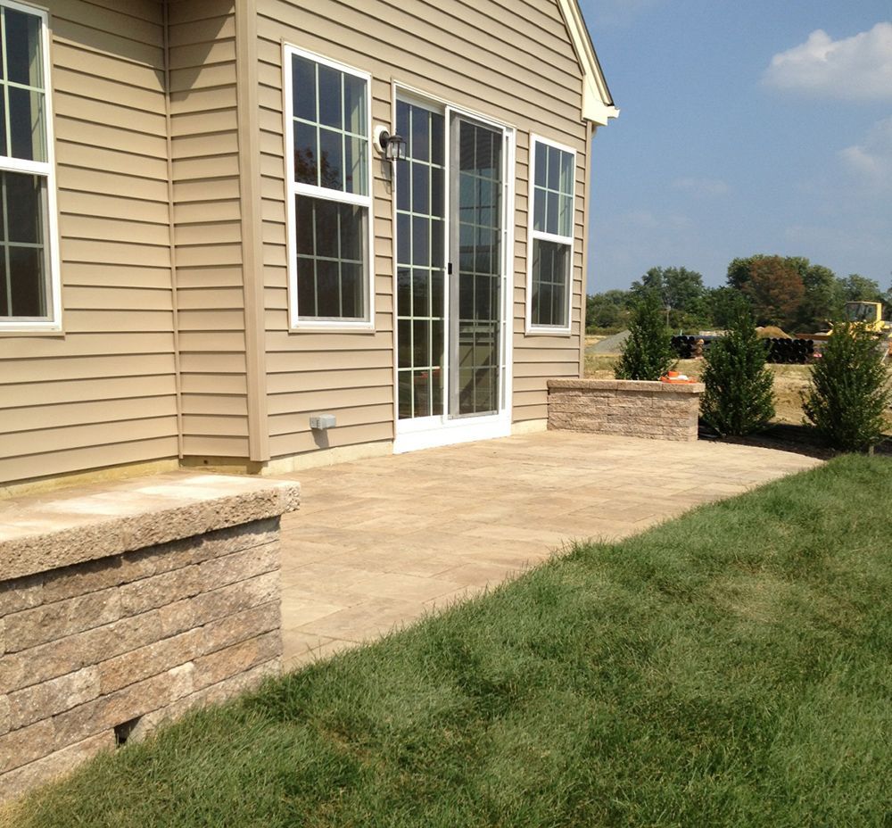 Backyard patio with sliding glass door and retaining walls; tan siding, green grass, and blue sky.