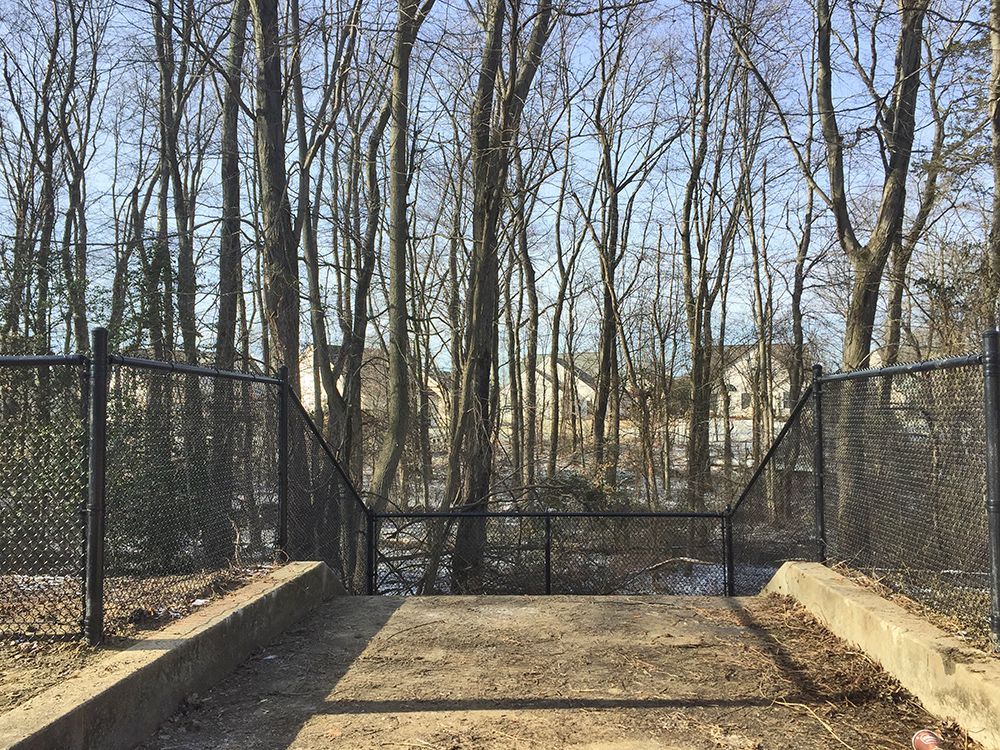 Chain-link fence with a concrete path leading to a wooded area. Trees line the background under a blue sky.