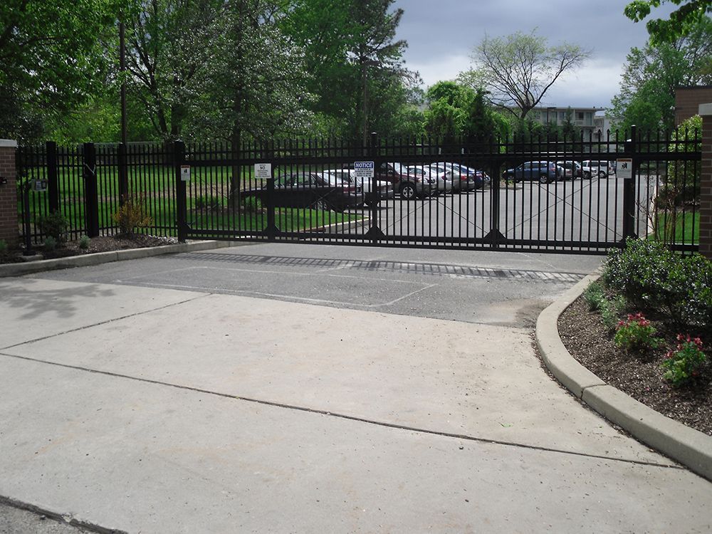 Black metal security gate at a paved entrance to a parking area, surrounded by brick and greenery.