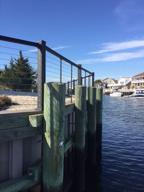 Dock with green pilings, black railing, blue water, and sky. Boats and houses visible in the background.