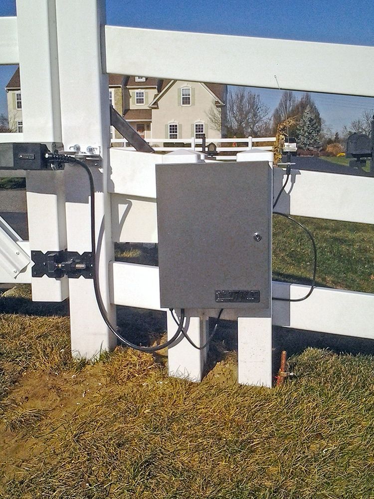 Gray metal box on a white fence post, likely for gate automation, with black wires and a grassy area.