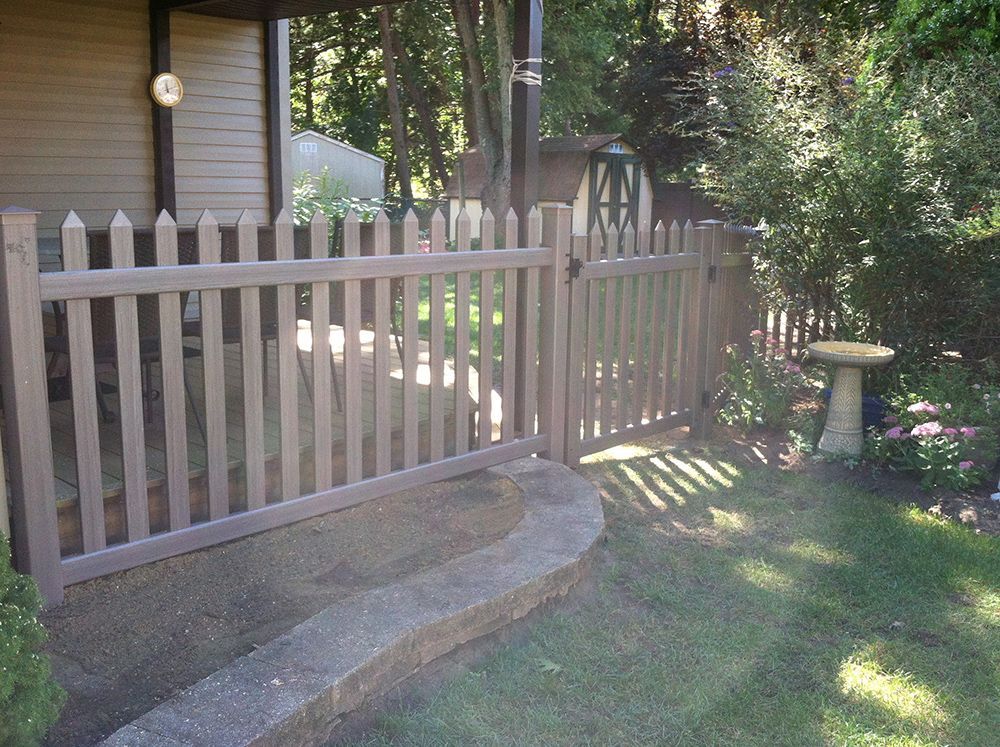 Brown picket fence with gate, bordering a grassy yard next to a building and garden.