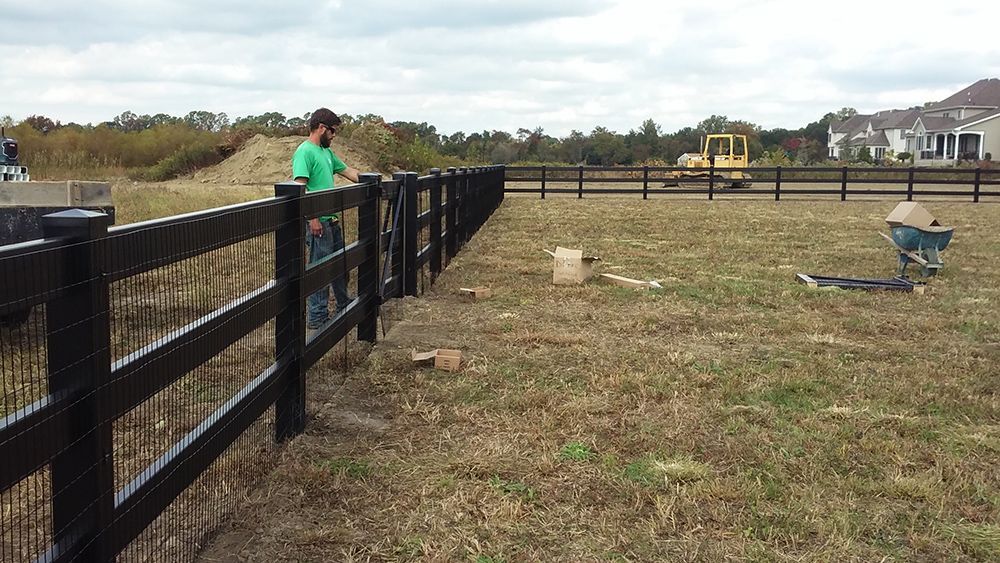 Man installing a black fence in a field; construction equipment and houses in the background.