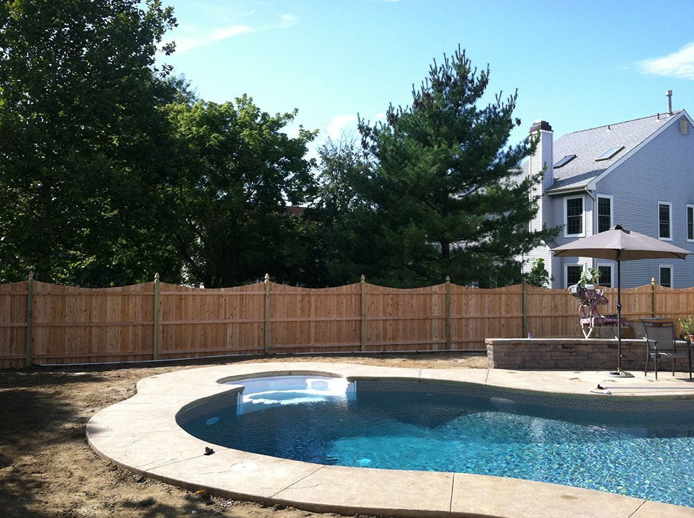 Backyard with a swimming pool, wooden fence, trees, and a house.