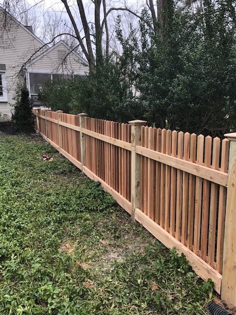 Wooden picket fence in a grassy yard, partially obscuring a house and greenery.