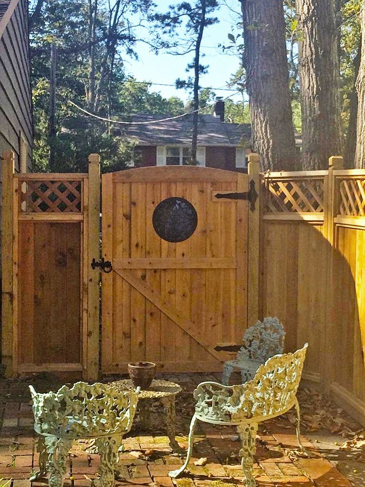 Wooden gate in a cedar fence with a round window, two ornate chairs, and a small table.
