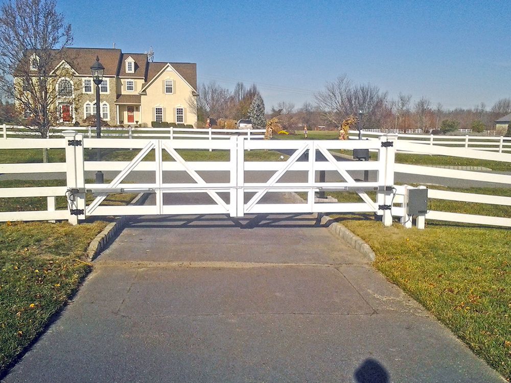 White gate and fence in front of a large house with a paved driveway on a sunny day.