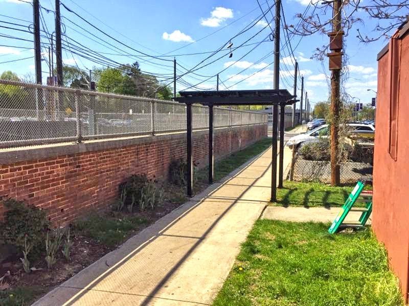A narrow brick and concrete path with a shaded pergola alongside a fence and train tracks.