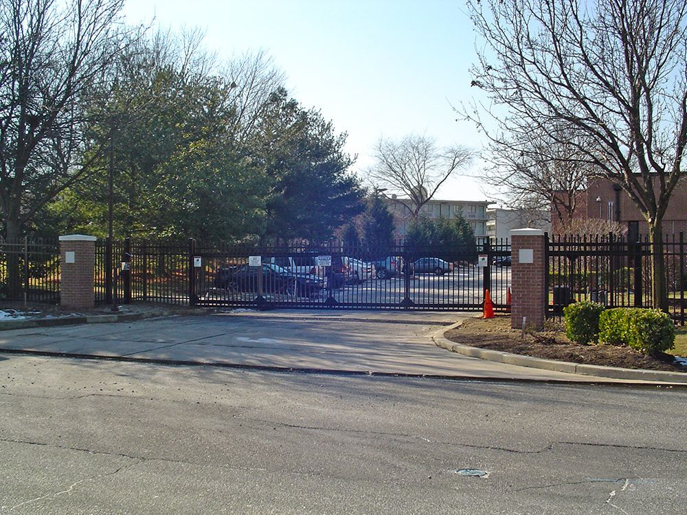 Gated entrance to a facility with brick pillars, black metal fence, and parked vehicles visible through the gate.