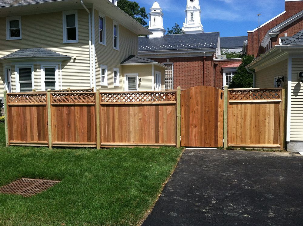 Wooden fence with gate, bordering a green lawn and driveway, with houses and church in the background.