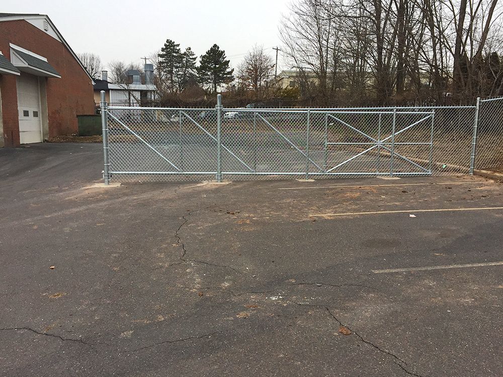 Chain-link fence with gate, in front of a brick building and trees, set in an asphalt parking lot.