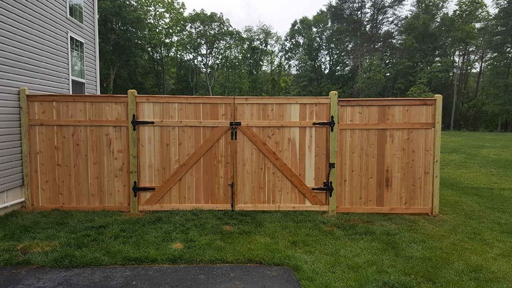 Wooden fence with double gate in a grassy backyard, adjacent to a house with trees in the background.