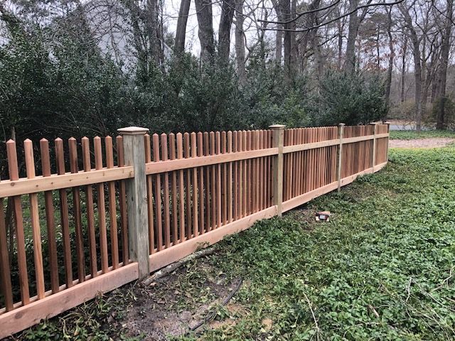 Wooden picket fence in a yard with bushes and trees in the background.