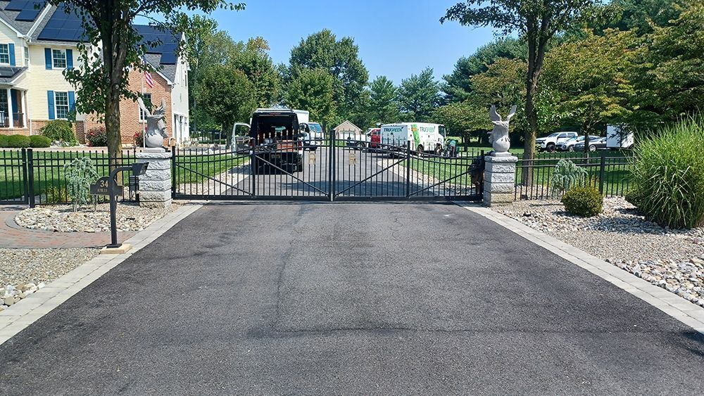 Gated driveway with asphalt leading to a house. Black iron gates are open, with vehicles parked behind them.