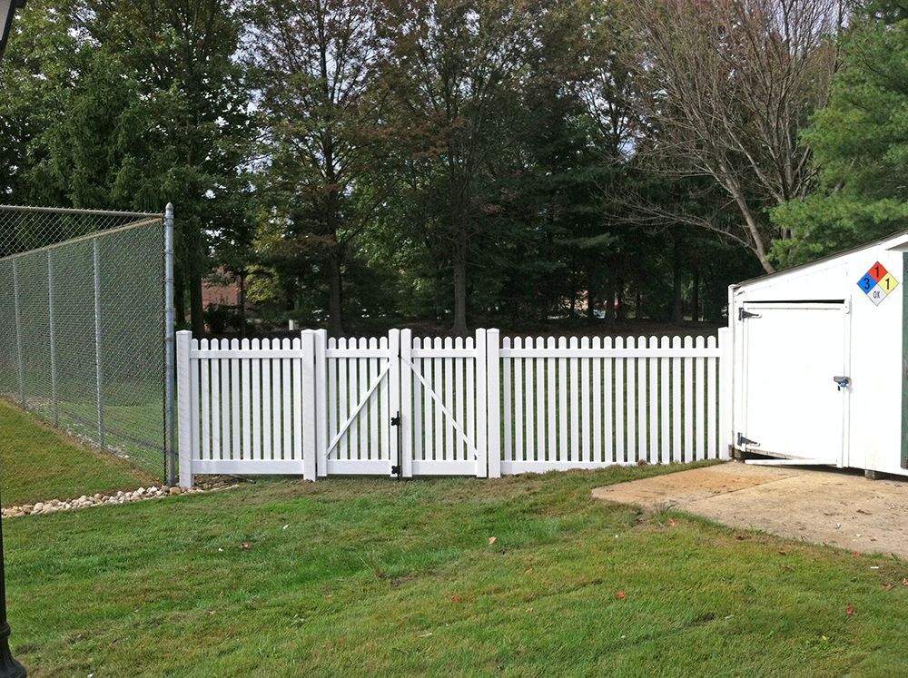 White picket fence with gate, grassy yard, and chain-link fence on the left side. Trees in the background.