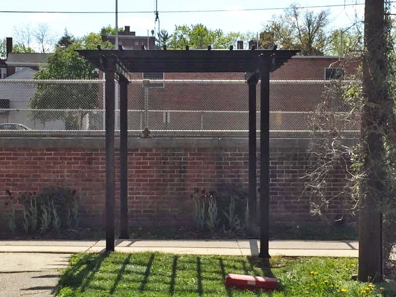Black pergola on grass with brick wall and chain-link fence in background. Sunny day, shadows cast.