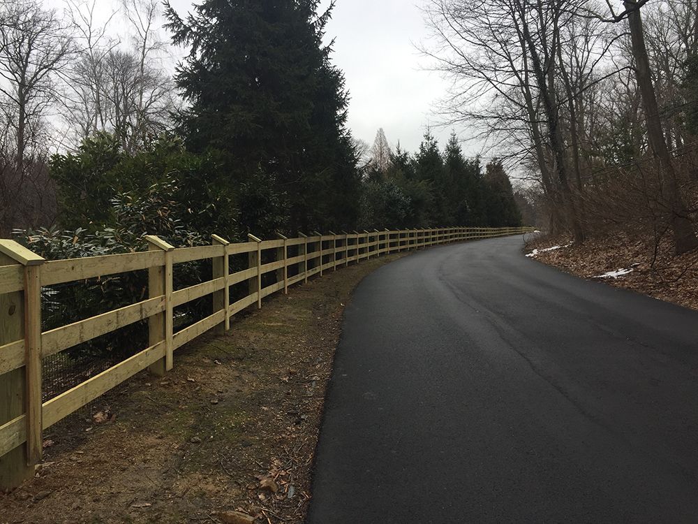 Wooden fence alongside a curved asphalt road, trees in the background, overcast day.