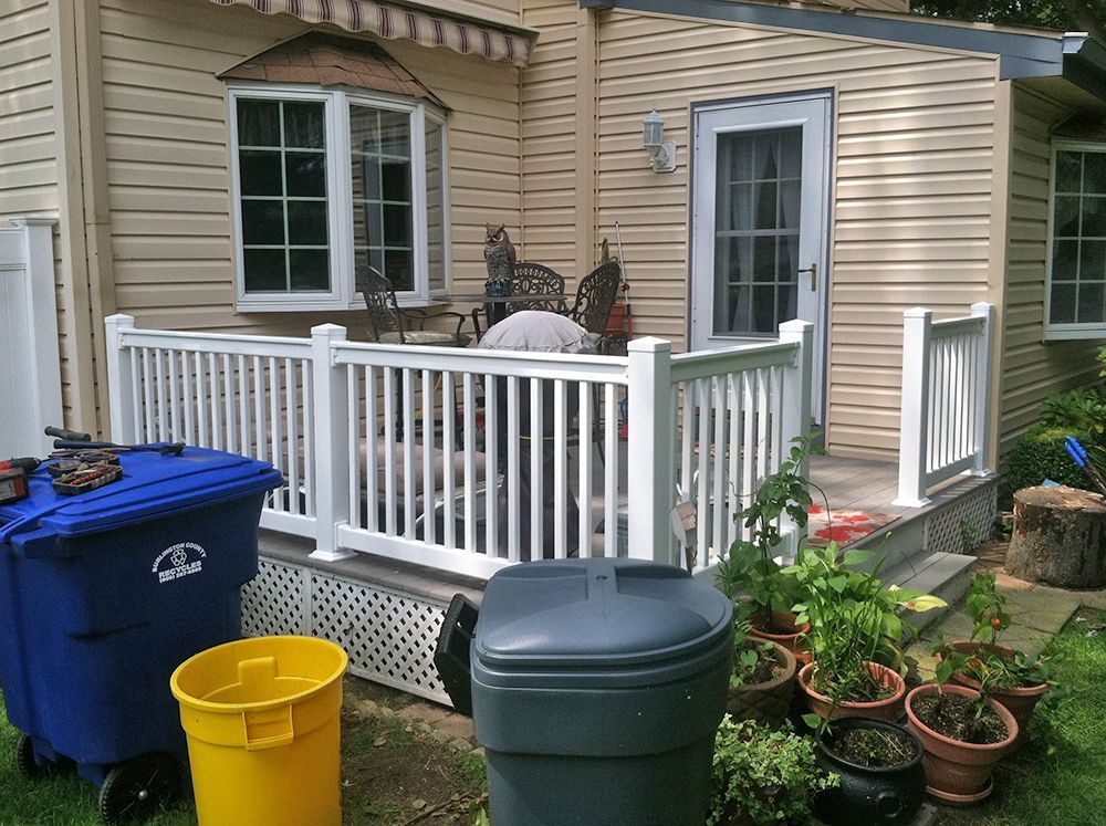 A small deck with white railing beside a light yellow house. A blue trash bin, yellow bucket, and compost bin sit nearby.
