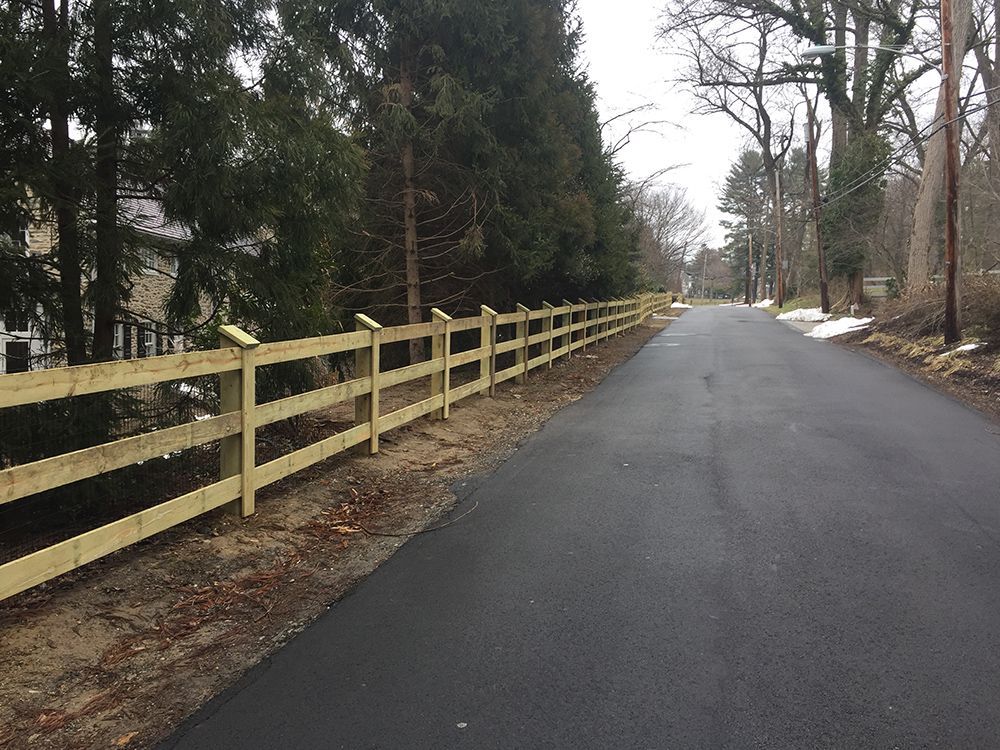 A newly paved road with a wooden fence alongside, bordered by trees on a cloudy day.