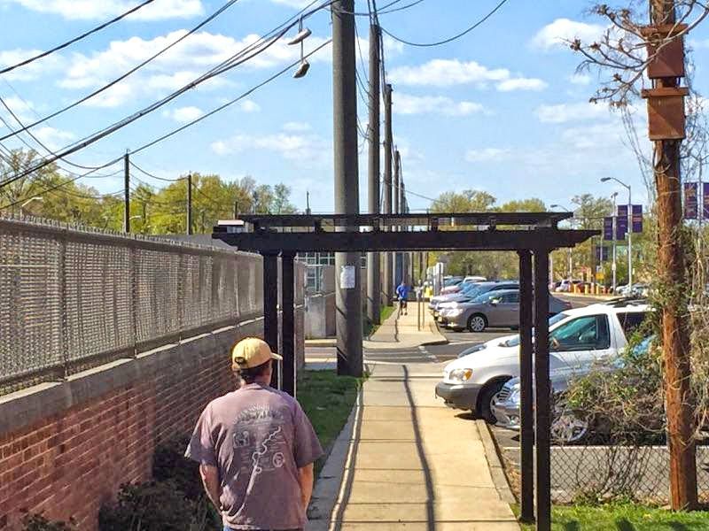 Person walks on sidewalk under a black archway next to a brick wall and parked cars. Power poles and blue sky visible.