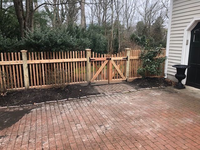 Wooden picket fence with gate in front of a house, set on a brick paved surface.
