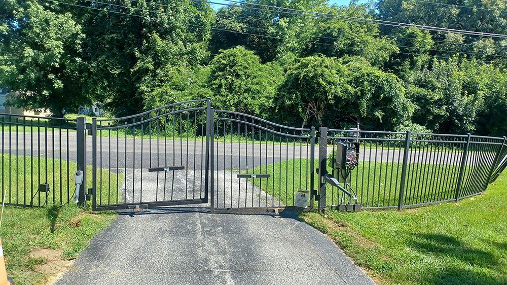 Black metal gate with curved top, protecting a driveway and a grassy yard, with trees in the background.