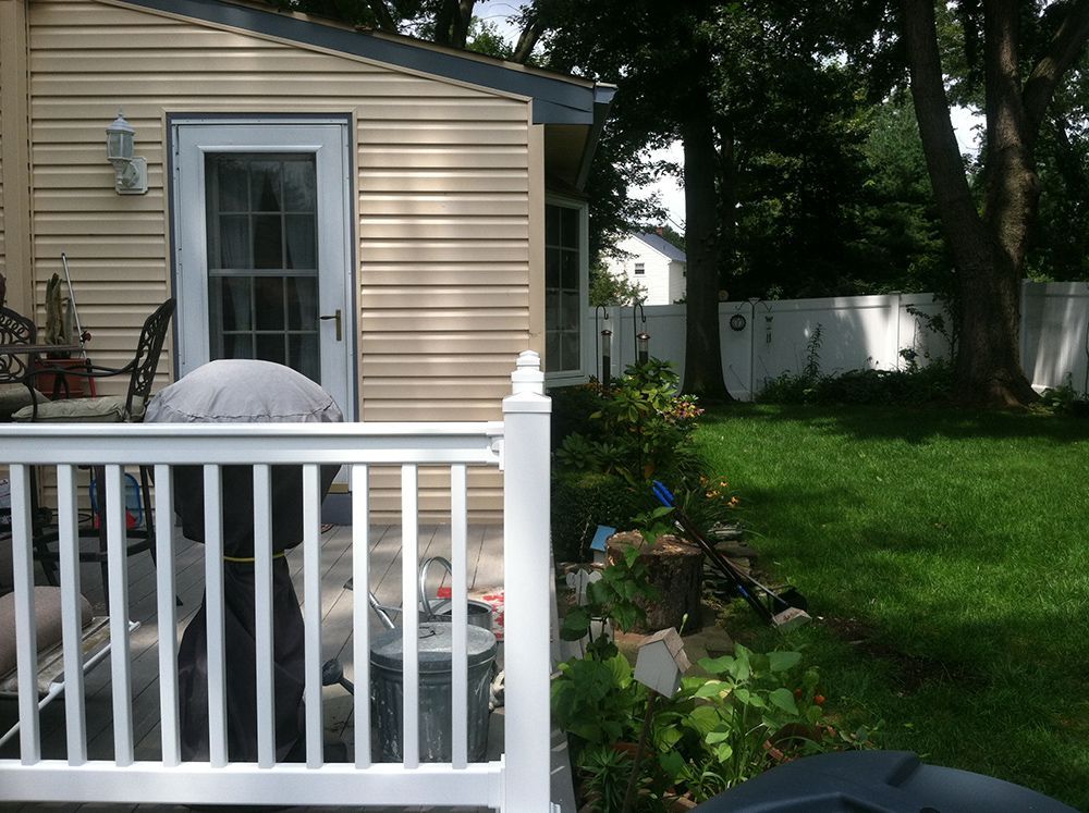 White porch railing and back door, beige house siding, green lawn, and tall trees.