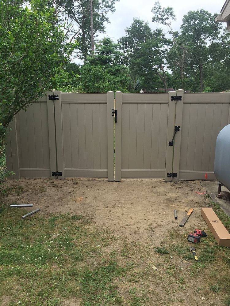 Tan vinyl fence with double gate, surrounded by dirt and grass, with tools on the ground.