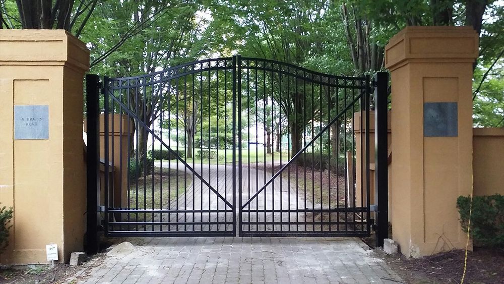 Black wrought-iron gate between two tan pillars, leading to a tree-lined driveway.