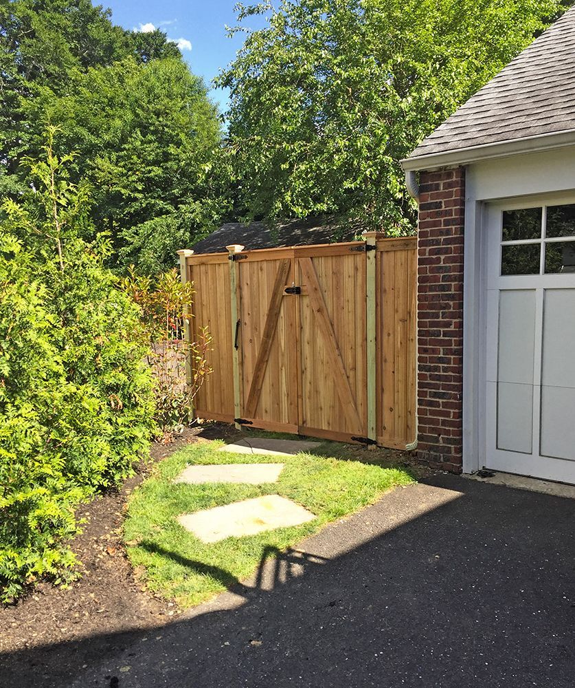 Wooden gate in a grassy area with stone pavers leading to a driveway next to a brick building.